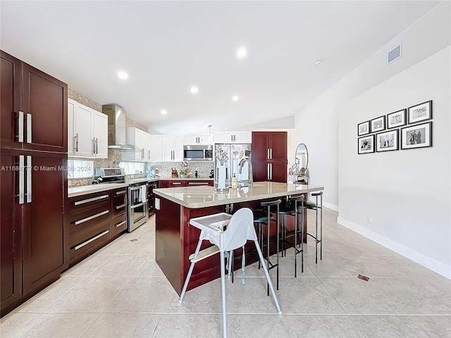 kitchen featuring wall chimney range hood, lofted ceiling, appliances with stainless steel finishes, a kitchen breakfast bar, and a kitchen island with sink