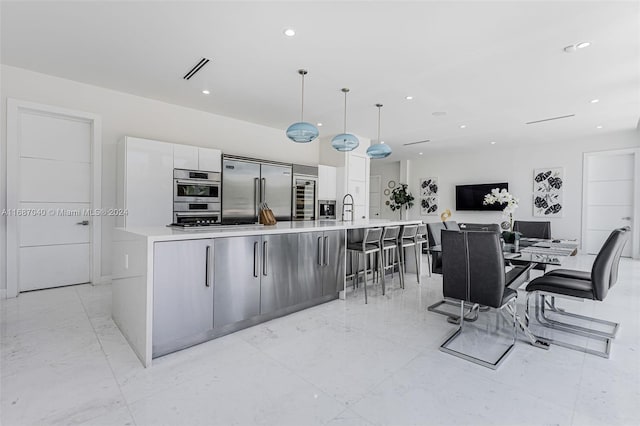 kitchen featuring a large island with sink, sink, appliances with stainless steel finishes, decorative light fixtures, and white cabinetry