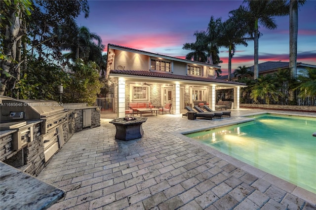 pool at dusk featuring a patio, exterior kitchen, an outdoor living space with a fire pit, and grilling area