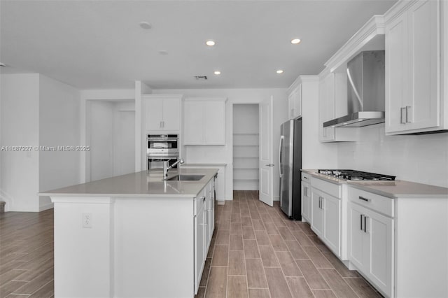 kitchen featuring wall chimney exhaust hood, an island with sink, stainless steel appliances, sink, and white cabinets