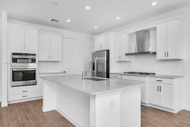 kitchen featuring wall chimney range hood, an island with sink, sink, white cabinetry, and appliances with stainless steel finishes