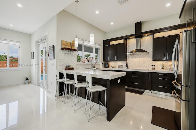 kitchen with wall chimney range hood, backsplash, a kitchen breakfast bar, stainless steel fridge, and decorative light fixtures