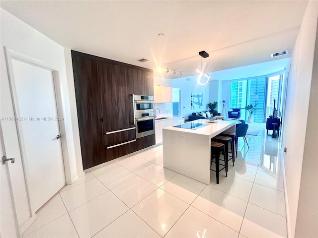 kitchen with a center island with sink, decorative light fixtures, dark brown cabinets, light tile patterned floors, and double oven