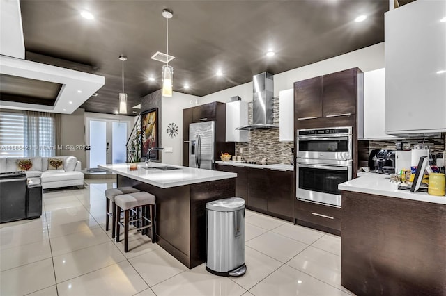 kitchen featuring wall chimney exhaust hood, a center island with sink, sink, decorative light fixtures, and appliances with stainless steel finishes