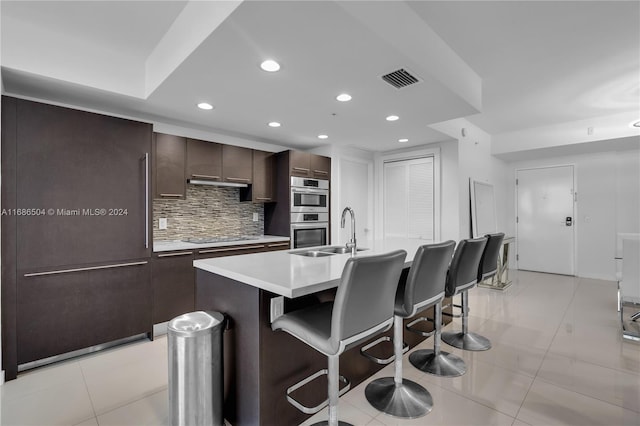 kitchen featuring sink, stainless steel double oven, backsplash, an island with sink, and a breakfast bar