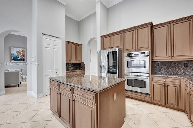 kitchen featuring light tile patterned floors, appliances with stainless steel finishes, dark stone countertops, a towering ceiling, and decorative backsplash