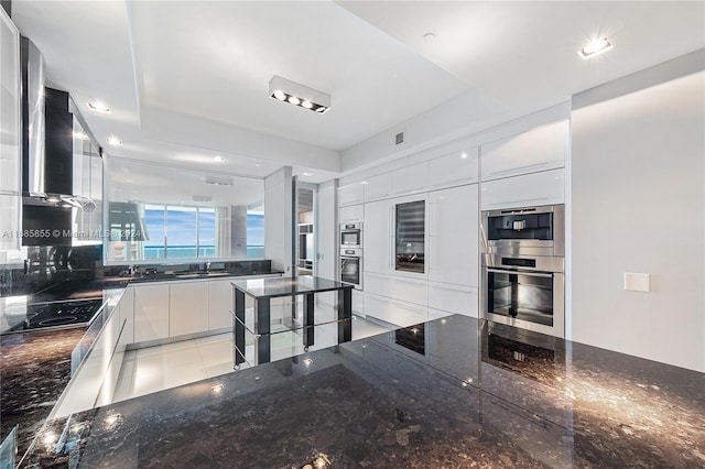 kitchen with wall chimney exhaust hood, dark stone countertops, stainless steel double oven, black electric cooktop, and white cabinetry