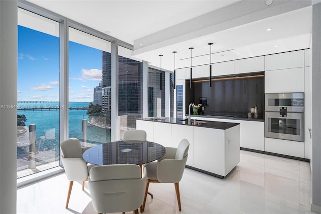 kitchen with stainless steel double oven, a center island with sink, pendant lighting, white cabinets, and a water view