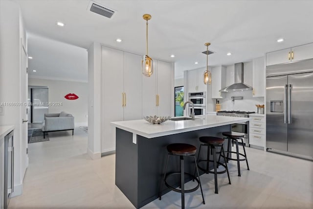 kitchen featuring wall chimney exhaust hood, high end appliances, white cabinetry, hanging light fixtures, and an island with sink