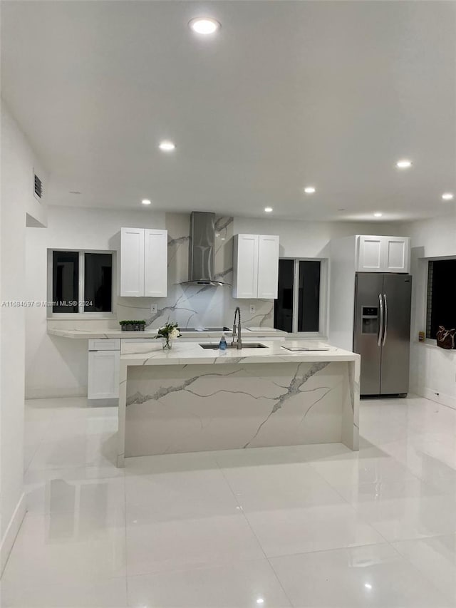 kitchen with stainless steel fridge, white cabinetry, light stone counters, and wall chimney range hood