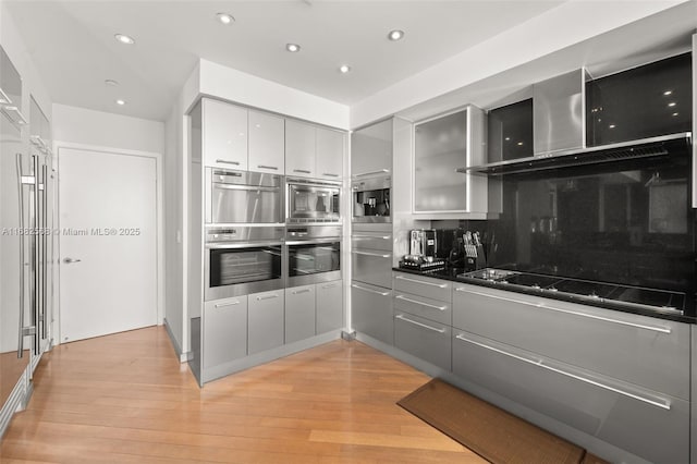 kitchen with wall chimney range hood, stainless steel appliances, light hardwood / wood-style floors, backsplash, and gray cabinetry