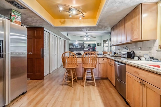 kitchen with appliances with stainless steel finishes, a kitchen breakfast bar, ceiling fan, a tray ceiling, and light hardwood / wood-style flooring