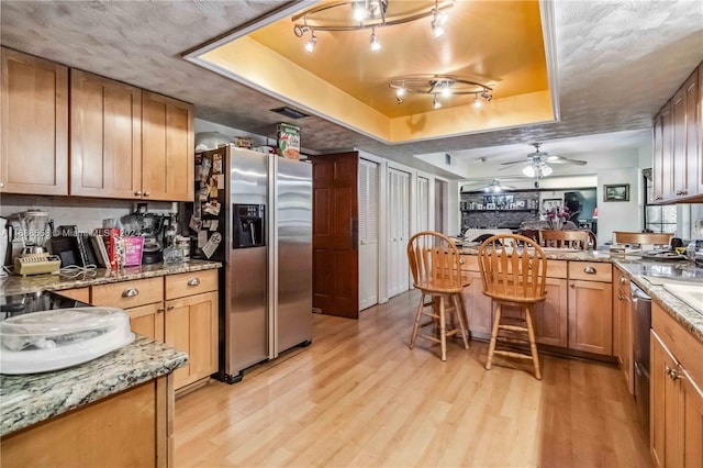 kitchen featuring light stone counters, a raised ceiling, stainless steel refrigerator with ice dispenser, ceiling fan, and light hardwood / wood-style flooring