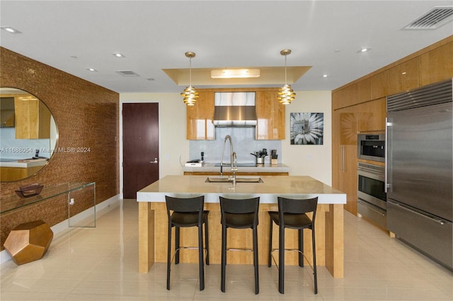 kitchen featuring stainless steel appliances, a kitchen island with sink, wall chimney exhaust hood, and pendant lighting