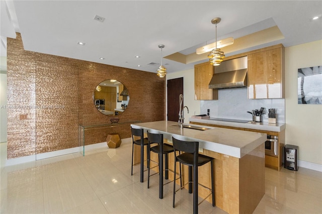 kitchen with a kitchen bar, wall chimney exhaust hood, sink, a tray ceiling, and decorative light fixtures