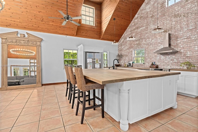 kitchen with butcher block counters, white cabinetry, high vaulted ceiling, wall chimney range hood, and pendant lighting