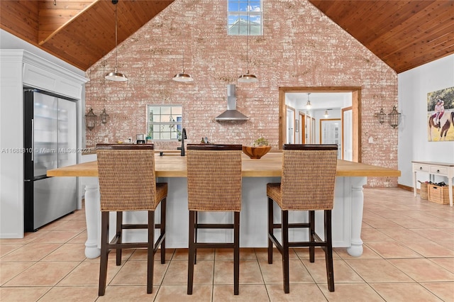 kitchen featuring wall chimney exhaust hood, high vaulted ceiling, a breakfast bar, decorative light fixtures, and stainless steel fridge