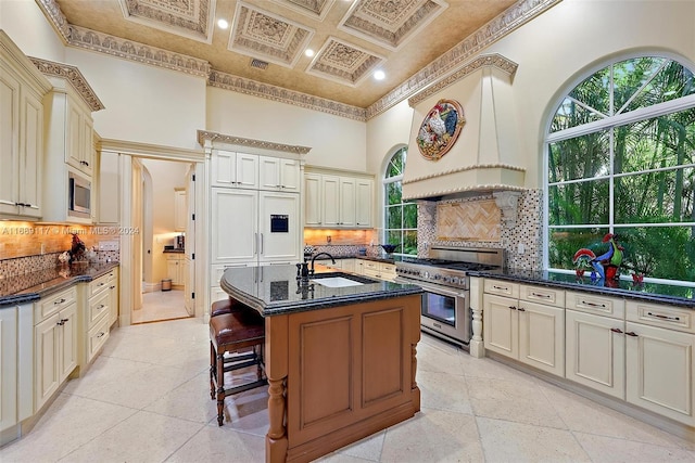 kitchen with coffered ceiling, appliances with stainless steel finishes, cream cabinetry, and a kitchen island with sink