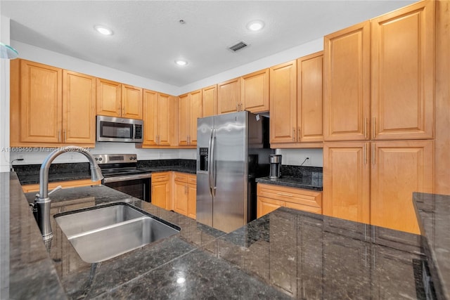 kitchen with stainless steel appliances, a textured ceiling, light brown cabinetry, sink, and dark stone countertops