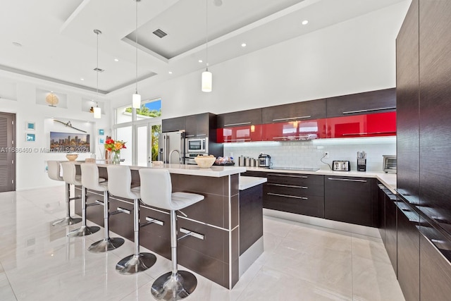 kitchen with stainless steel appliances, decorative backsplash, hanging light fixtures, an island with sink, and a high ceiling