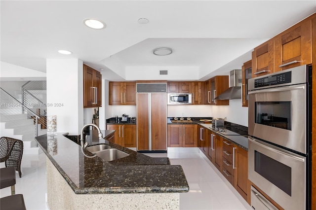 kitchen with built in appliances, wall chimney exhaust hood, sink, light tile patterned floors, and dark stone countertops