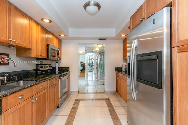 kitchen with stainless steel appliances, light tile patterned flooring, sink, dark stone counters, and a tray ceiling