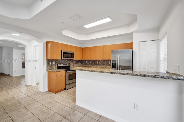 kitchen with stainless steel appliances, kitchen peninsula, light tile patterned flooring, backsplash, and dark stone countertops