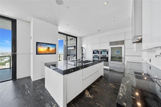 kitchen with floor to ceiling windows, sink, white cabinets, a kitchen island with sink, and wall chimney range hood