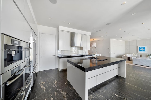 kitchen featuring sink, an island with sink, white cabinets, oven, and wall chimney range hood