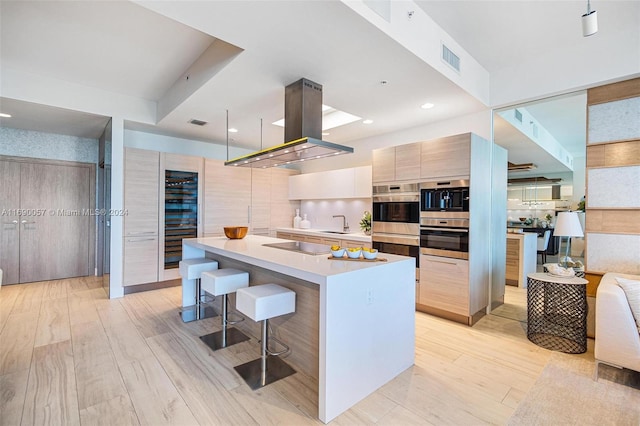 kitchen featuring double oven, island exhaust hood, a kitchen bar, a kitchen island, and light wood-type flooring