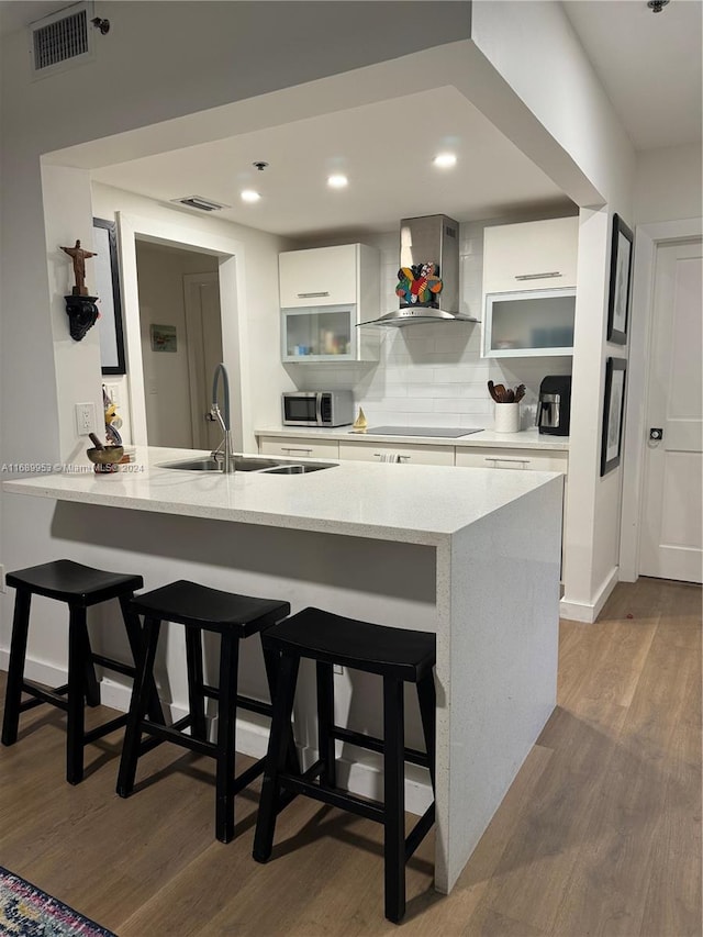 kitchen featuring kitchen peninsula, wall chimney exhaust hood, a kitchen breakfast bar, white cabinetry, and light hardwood / wood-style flooring