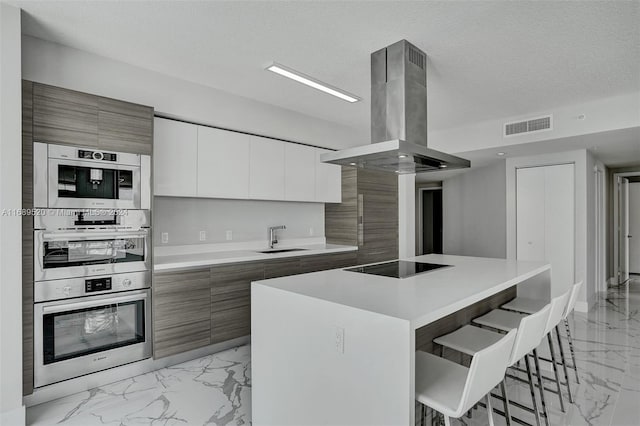 kitchen featuring black electric cooktop, island range hood, sink, a kitchen breakfast bar, and white cabinetry