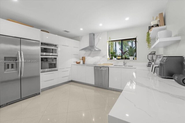 kitchen with sink, wall chimney exhaust hood, light stone countertops, white cabinetry, and stainless steel appliances