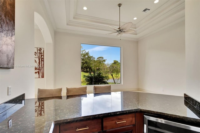 kitchen with beverage cooler, dark stone counters, ornamental molding, ceiling fan, and a tray ceiling