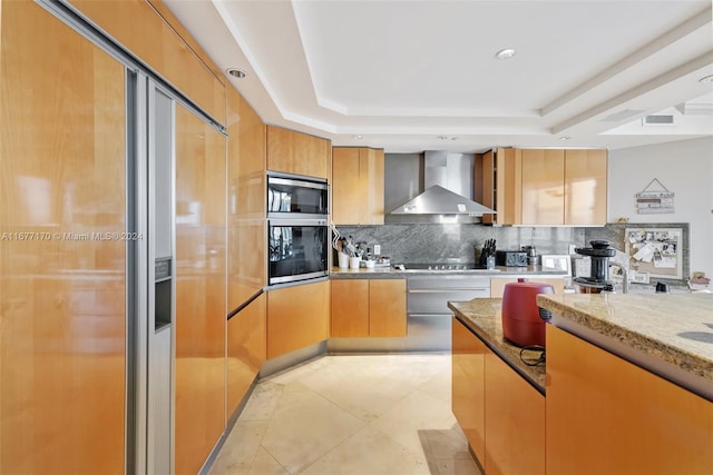 kitchen featuring backsplash, wall chimney range hood, a tray ceiling, and light stone counters