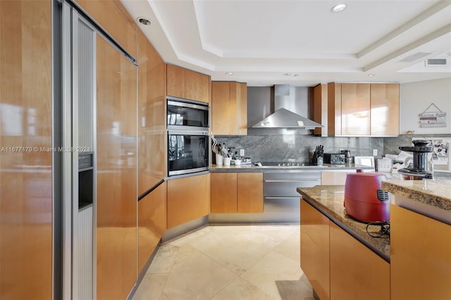 kitchen featuring stainless steel appliances, decorative backsplash, wall chimney range hood, a tray ceiling, and dark stone countertops