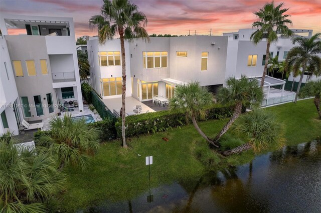 back house at dusk with a water view, a balcony, a patio area, a lawn, and a pool