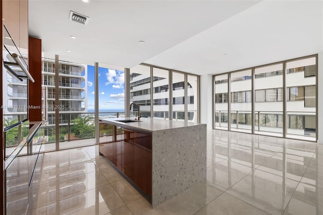 kitchen with sink, light tile patterned flooring, expansive windows, an island with sink, and light stone countertops