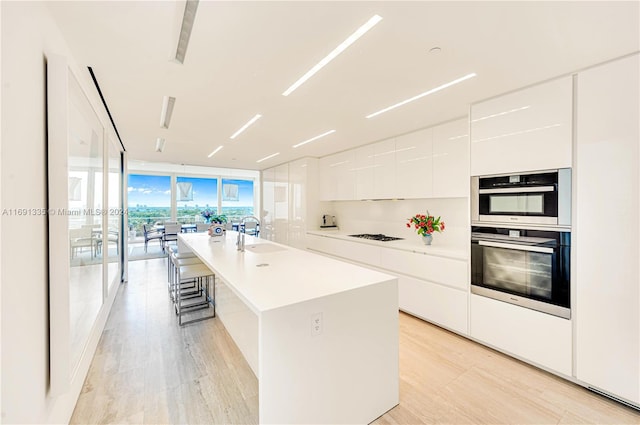 kitchen featuring sink, a wall of windows, an island with sink, light hardwood / wood-style flooring, and white cabinets