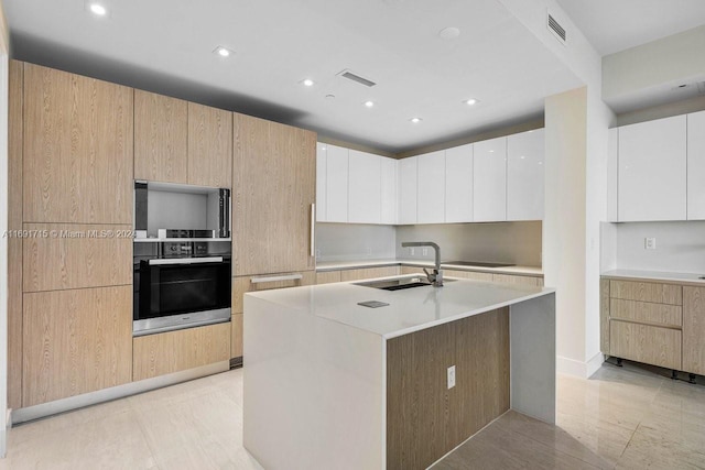 kitchen with double oven, a kitchen island with sink, sink, light tile patterned floors, and white cabinetry