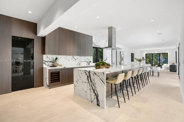 kitchen featuring island exhaust hood, light stone counters, wine cooler, a large island, and decorative backsplash