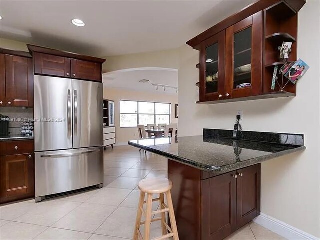 kitchen with light tile patterned flooring, stainless steel refrigerator, a breakfast bar, kitchen peninsula, and dark stone countertops