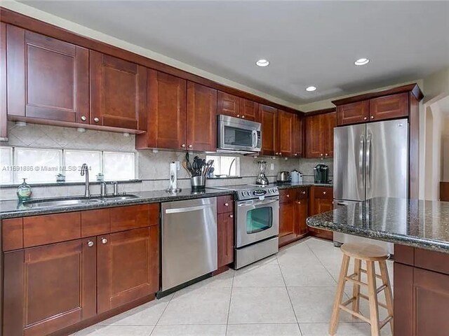 kitchen with stainless steel appliances, dark stone counters, light tile patterned floors, sink, and a breakfast bar area