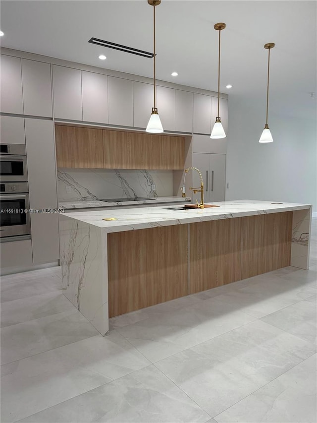 kitchen with an island with sink, hanging light fixtures, sink, and light stone counters