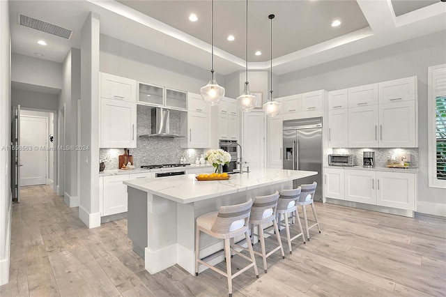 kitchen with stainless steel appliances, a center island with sink, white cabinetry, light stone counters, and wall chimney exhaust hood