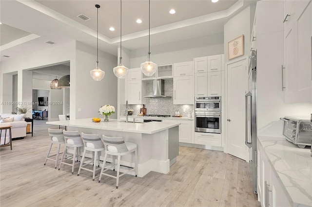 kitchen with stainless steel appliances, light wood-type flooring, white cabinets, a tray ceiling, and wall chimney exhaust hood