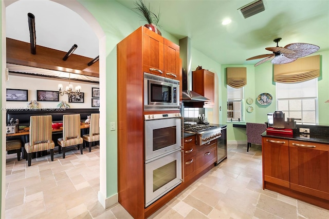 kitchen featuring wall chimney range hood, appliances with stainless steel finishes, decorative light fixtures, and ceiling fan with notable chandelier