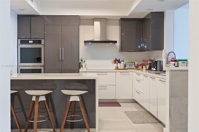 kitchen featuring white cabinetry, stovetop, wall chimney exhaust hood, a breakfast bar area, and stainless steel double oven