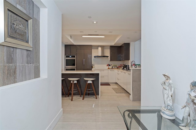 kitchen with stainless steel double oven, white cabinetry, wall chimney range hood, dark brown cabinetry, and a breakfast bar