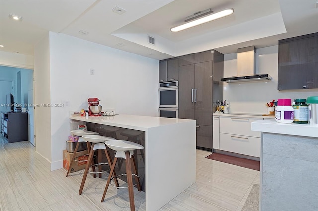 kitchen featuring white cabinets, a breakfast bar area, stainless steel double oven, a raised ceiling, and wall chimney range hood